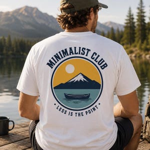 Könnte beinhalten: Ein weißes Kurzarm-T-Shirt mit einer kreisförmigen Grafik auf der Rückseite. Das Design zeigt einen dunkelblauen Berg, eine gelbe Sonne und ein dunkelblaues Kanu auf hellblauem Wasser. Der Text lautet MINIMALIST CLUB und LESS IS THE POINT.