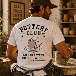 Könnte beinhalten: Ein weißes kurzärmeliges T-Shirt mit einem Grafikdruck auf der Rückseite. Das Design zeigt den Text POTTERY CLUB und LIFE IS BETTER ON THE WHEEL in Schwarz und Olivgrün sowie eine Illustration von Händen, die einen Tontopf formen.