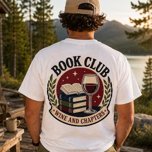 Könnte beinhalten: Ein weißes T-Shirt mit einem Grafikdruck auf der Rückseite, der den Text BOOK CLUB und WINE AND CHAPTERS zeigt. Das Motiv besteht aus einem Stapel von drei Büchern, einem aufgeschlagenen Buch und einem Weinglas, umrahmt von grünen Lorbeerblättern.