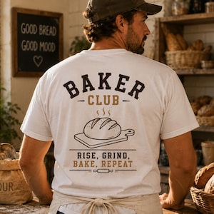 Könnte beinhalten: Ein weißes T-Shirt mit einer dunkelbraunen Grafik auf der Rückseite. Das Design zeigt den Schriftzug "BAKER CLUB" über einer Illustration eines Brotlaibs auf einem Schneidebrett sowie den Text "RISE, GRIND, BAKE, REPEAT" und ein Nudelholz-Symbol.