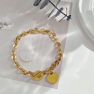 Può includere: Un bracciale a catena color oro con chiusura a levetta e un ciondolo a forma di cuore. Il ciondolo ha un testo inciso. Il bracciale è su una superficie bianca con un rametto di verde.