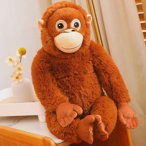 May include: A plush orangutan toy with brown fur, large eyes, and a smiling expression. The stuffed animal is sitting with its arms and legs extended. The toy is designed to resemble a real orangutan.