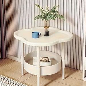 Puede incluir: Una mesa auxiliar blanca en forma de nube con un estante inferior. Una taza azul y un jarrón de cristal con vegetación están sobre la mesa. La mesa tiene un diseño minimalista y es adecuada para una sala de estar o dormitorio.