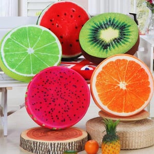 Könnte beinhalten: Eine Sammlung runder, fruchtförmiger Kissen, darunter eine Wassermelone, Kiwi, Limette, Drachenfrucht und Orange. Die Kissen sind farbenfroh gestaltet und ahmen die Querschnitte der Früchte nach. Dekorative Heimdekoration.