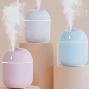 Op de afbeelding: Drie pastelkleurige, eivormige etherische olie-diffusers. De diffusers zijn lichtroze, lichtblauw en wit, elk met een gloeiende witte horizontale streep en een klein lampje bovenop. Elke diffuser geeft een stroom witte damp af.
