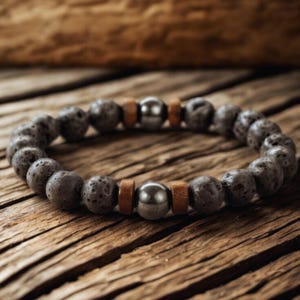 Op de afbeelding: Een armband met donkergrijze lavasteen kralen, metallic grijze kralen en kleine houten spacers. De armband wordt gepresenteerd op een rustiek houten oppervlak, dat de natuurlijke texturen en aardetinten benadrukt. De omtrek is ongeveer 20 cm.