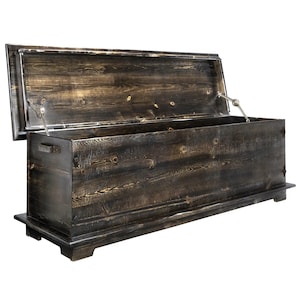 May include: A large wooden chest with a dark, distressed finish and an open lid. The chest is rectangular with a hinged lid and metal hardware. It has a handle on the side and sits on a low base. The wood grain is visible throughout.
