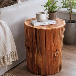 May include: A cylindrical wooden side table made from a natural tree stump with visible grain, knots, and vertical cracks. A white ceramic mug and a book titled The Kinfolk Home are placed on the flat, circular top surface of the wooden stump.