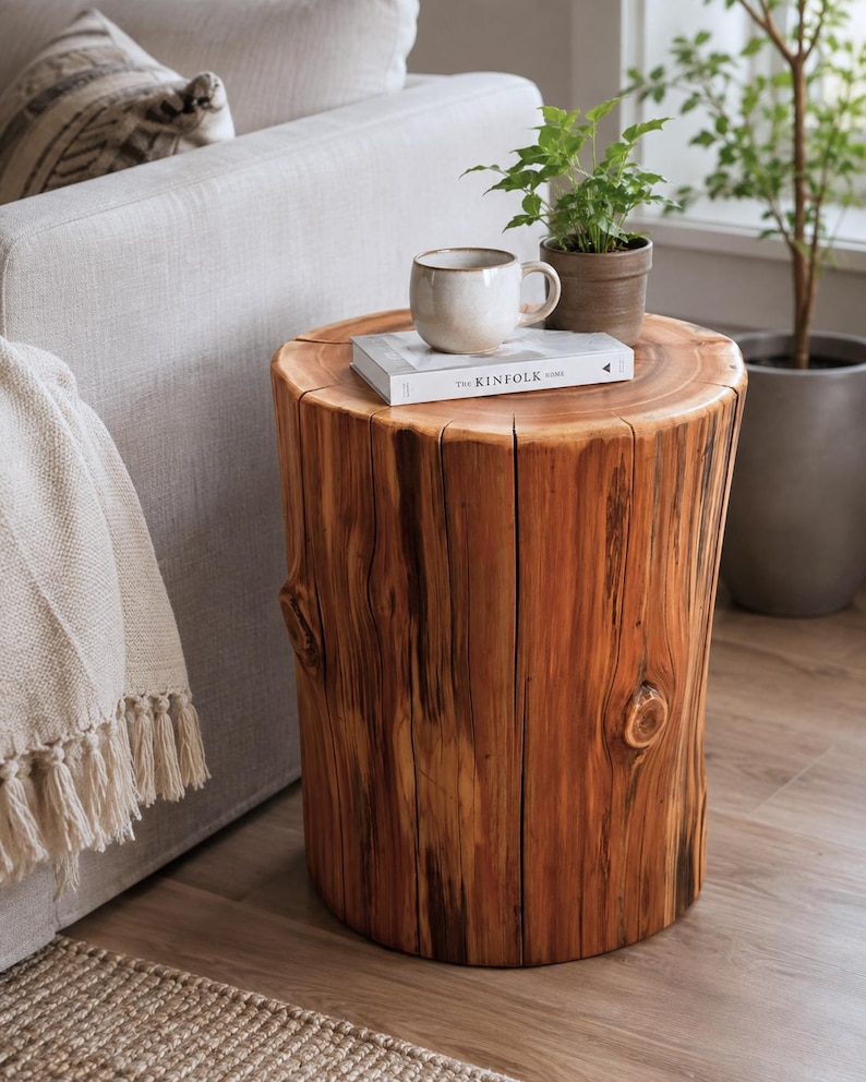 Pu&ograve; includere: Un tavolino cilindrico in legno ricavato da un ceppo d'albero naturale con venature, nodi e crepe verticali visibili. Una tazza in ceramica bianca e un libro intitolato The Kinfolk Home sono appoggiati sulla superficie circolare superiore.