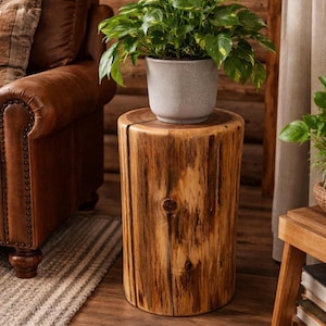 May include: A cylindrical wooden side table with a natural wood grain finish. A potted plant with green leaves sits on top. The table is approximately 20 inches tall and 15 inches in diameter.