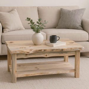 May include: A rustic, light-colored wooden coffee table with a lower shelf. The table has a natural, unfinished look. A small, round, cream-colored vase with greenery, a black mug, and a book sit on top. A beige sofa and pillows are in the background.