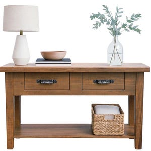 May include: A wooden console table with two drawers, a lower shelf, and a woven basket. A white lamp with a beige shade, a wooden bowl, books, and a glass vase with greenery are on top. The table is brown.