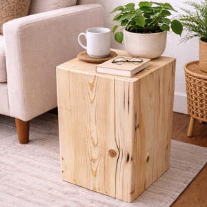 May include: A square, light-colored wooden block side table featuring a natural grain pattern with visible knots and vertical cracks. The top surface holds a white ceramic mug on a round coaster, a closed beige book, and metal-rimmed glasses.