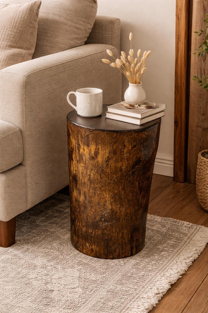 Puede incluir: Una mesa auxiliar cil&iacute;ndrica hecha de un toc&oacute;n de &aacute;rbol natural de color marr&oacute;n oscuro con una superficie superior plana y lisa. La mesa sostiene una taza de cer&aacute;mica blanca, un peque&ntilde;o jarr&oacute;n blanco con hierbas secas y dos libros apilados.