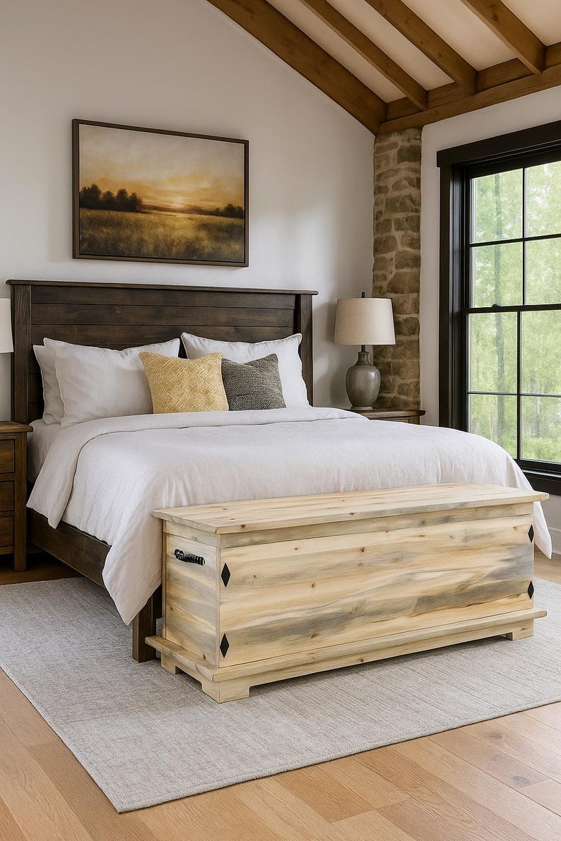 May include: A bedroom with a dark wooden bed, white bedding, and decorative pillows. A light wood chest is at the foot of the bed. A framed landscape painting hangs above the bed. The room has a rustic feel.