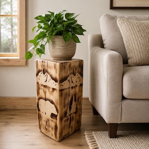 May include: A square wooden side table featuring a scorched finish with carved mountain and bear silhouettes. A light-colored, textured ceramic pot sits on top, containing a vibrant green pothos plant with trailing leaves and stems.