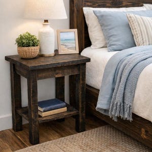 May include: A dark brown wooden nightstand with a lower shelf. A lamp with a white shade and a small potted plant sit on top. A framed picture and two books are also visible. The nightstand is next to a bed with blue pillows and a blue blanket.