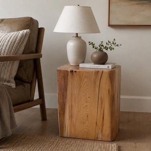May include: A square wooden side table with a natural grain finish, topped with a white ceramic table lamp and a small brown vase containing green foliage. A book titled This Is Home rests on the surface of the wooden block table.