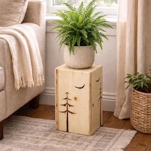 May include: A square wooden side table featuring a light natural finish with wood-burned illustrations of pine trees and a bird on the front panel. A speckled light gray ceramic pot containing a vibrant green fern sits on the flat top surface.