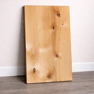 May include: A rectangular wooden cutting board with a light brown color and visible wood grain. The board has several knots and is leaning against a white wall. The floor is a light gray color.