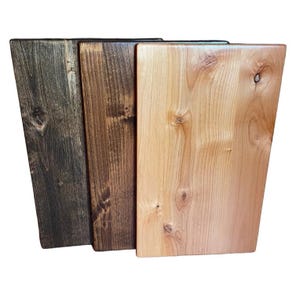 May include: Three wooden cutting boards in varying shades of brown. The boards are rectangular with a trapezoidal shape. The wood grain is visible, and the boards have a smooth finish. The colors range from dark brown to light tan.
