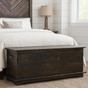 May include: A dark brown wooden storage chest, rectangular with a flat top, metal handles, and placed at the foot of a bed. The headboard is made of dark wood. The chest is approximately 150 cm long.