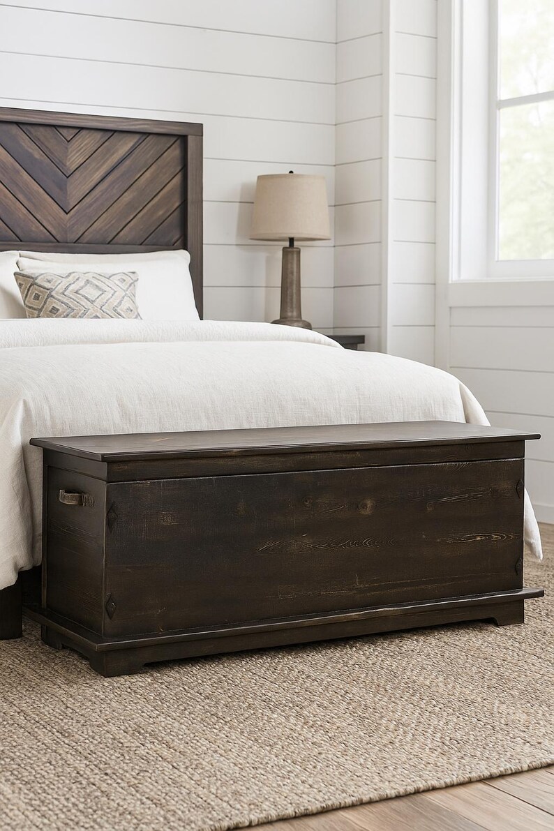 May include: A dark brown wooden storage chest, rectangular with a flat top, metal handles, and placed at the foot of a bed. The headboard is made of dark wood. The chest is approximately 150 cm long.