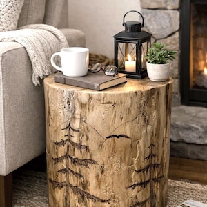 May include: A cylindrical wooden log side table featuring a wood-burned design of two pine trees, a mountain range, and a bird in flight. The natural light-colored wood grain is visible, and the top surface holds a white mug, a book, and glasses.