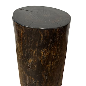 Puede incluir: Un taburete cil&iacute;ndrico de madera con un acabado oscuro te&ntilde;ido. La superficie superior muestra patrones de veta de madera natural y una grieta visible en el centro. Los lados presentan una apariencia rugosa y texturizada en tonos marrones.