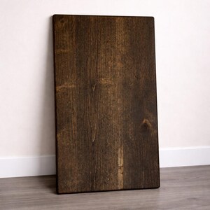 May include: A rectangular, dark brown wooden board with visible wood grain and a knot. The board is leaning against a white wall and rests on a gray floor. The wood has a rich, warm tone.