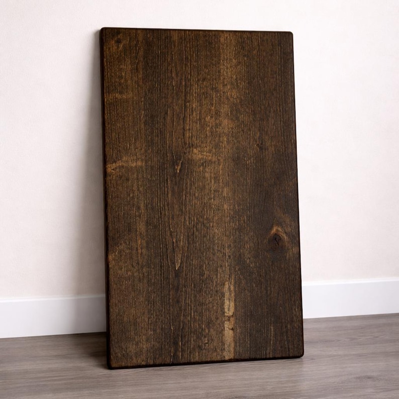 May include: A rectangular, dark brown wooden board with visible wood grain and a knot. The board is leaning against a white wall and rests on a gray floor. The wood has a rich, warm tone.