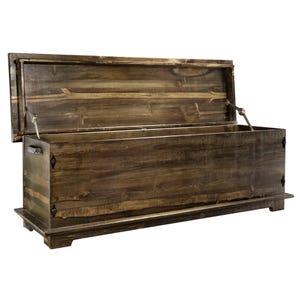 May include: A large, open wooden chest with a hinged lid. The chest is made of dark brown wood with visible grain and is rectangular in shape. The lid is propped open, revealing the interior. The chest sits on a low base and has metal hardware.