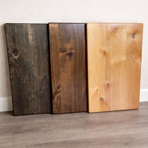 May include: Three wooden cutting boards in varying shades of brown and tan. The boards are rectangular with rounded corners. The wood grain is visible, and the boards are arranged vertically against a white wall and gray floor.