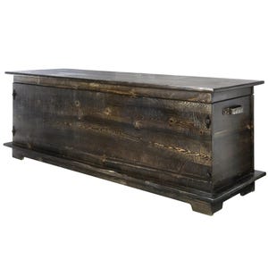 May include: A dark stained wooden storage chest. The rectangular chest has a flat top, a base, and metal handles. The wood grain is visible, and the chest appears handcrafted. The chest is approximately 150 cm long.