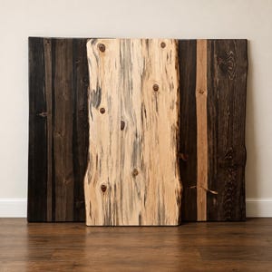 May include: A rectangular wooden board with a natural light-colored wood grain featuring dark knots and streaks, positioned in front of two dark brown, vertically oriented wooden planks with visible grain patterns and knots.