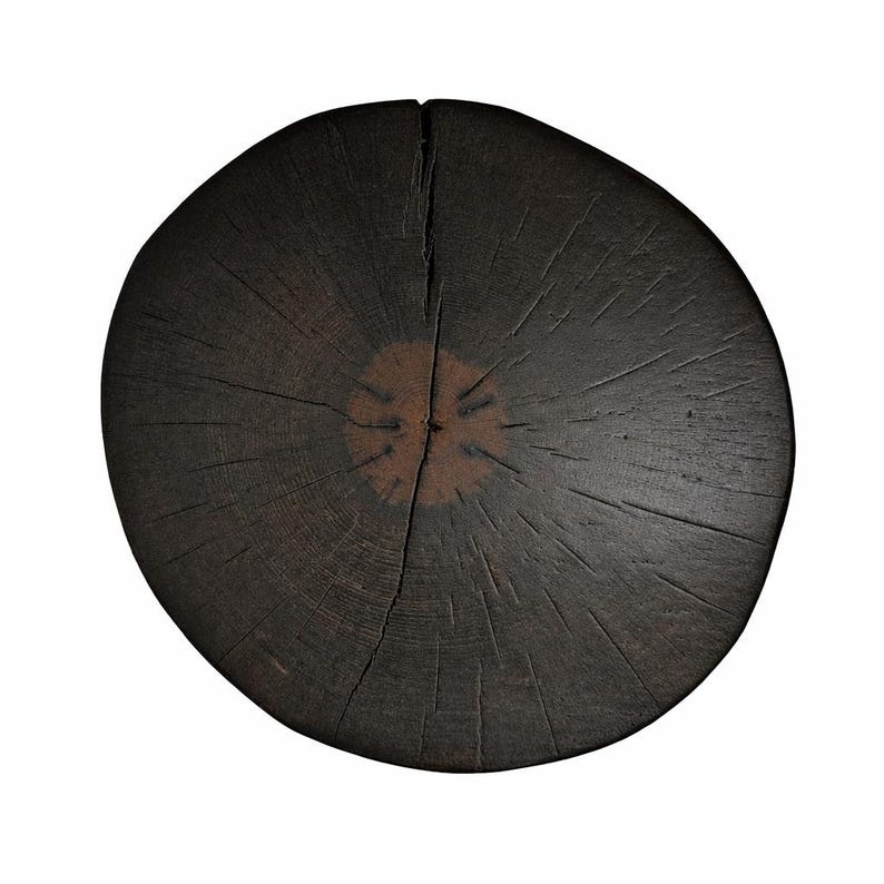 Puede incluir: Una rodaja de madera circular con un acabado marr&oacute;n oscuro carbonizado. La superficie muestra un anillo central prominente en un tono marr&oacute;n m&aacute;s claro, rodeado de profundas vetas radiantes y una grieta vertical que va del centro al borde.