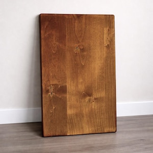 May include: A rectangular wooden board with a rich, warm brown stain. The wood grain is visible, and the board is leaning against a white wall. The edges are clean and the overall appearance is smooth.