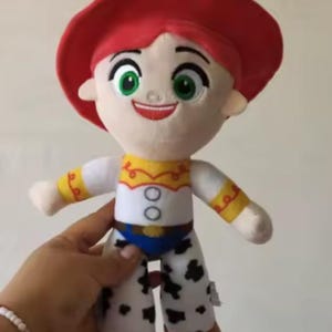 May include: A plush doll of the character Jessie from Toy Story. The doll has a red hat, red hair, and a white shirt with yellow accents. Her pants are white with black spots, and she has brown boots. The doll is held by a person.