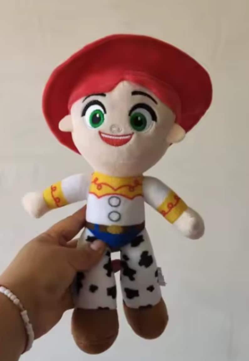 May include: A plush doll of the character Jessie from Toy Story. The doll has a red hat, red hair, and a white shirt with yellow accents. Her pants are white with black spots, and she has brown boots. The doll is held by a person.
