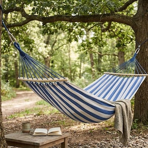 May include: A blue and white striped hammock hangs between two trees in a forest. A small wooden table holds a mug and an open book. A tan blanket is draped over the hammock.