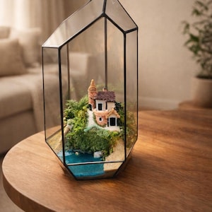 Peut inclure: Un terrarium géométrique en verre avec un paysage miniature. À l'intérieur, une petite maison avec un toit rouge et un phare se trouve sur une colline verte et moussue, à côté d'un plan d'eau bleu. Le terrarium est sur une table ronde en bois.