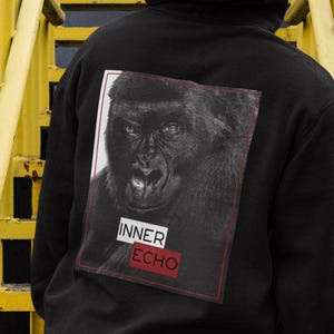 Op de afbeelding: Zwarte hoodie met een gorilla-afbeelding op de rug. De afbeelding is een zwart-wit afbeelding van het gezicht van een gorilla met de tekst "INNER ECHO" in een rode kader. De persoon staat op gele trappen.