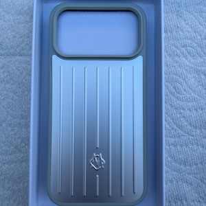 May include: A rectangular silver phone case with a vertical ribbed texture and a gray border frame. The case features a small, embossed logo in the center and a large rectangular cutout at the top, all presented inside a white rectangular box.