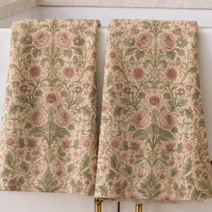May include: Two rectangular kitchen towels featuring a repeating botanical pattern of dusty pink roses and intricate sage green foliage on a cream-colored background. The towels are draped over the edge of a white farmhouse sink in a kitchen.