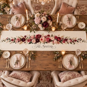 May include: A cream-colored linen table runner featuring a central floral arrangement of deep red, pink, and white roses with green foliage. The text "The SMITHS" is printed in black script in the center. The runner is set on a wooden table.