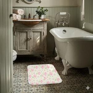 以下が含まれることがあります： 淡いクリーム色の背景にピンクの花と緑の葉のフローラルパターンが施された長方形のバスマット。バスマットは、爪足のバスタブと木製の洗面化粧台のあるバスルームにあります。