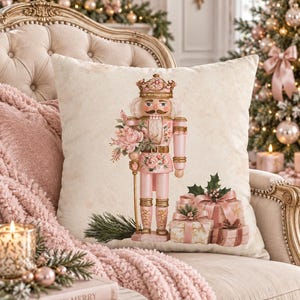 May include: A square throw pillow featuring a cream-colored background with a central illustration of a nutcracker wearing a crown and pink uniform, holding a bouquet of pink flowers, accompanied by a stack of wrapped gifts with pink ribbons.