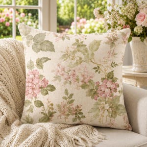 May include: Square decorative pillow with a floral pattern featuring pink hydrangeas and green leaves on a cream background. The pillow is placed on a beige sofa with a knitted throw, creating a cozy, inviting aesthetic.