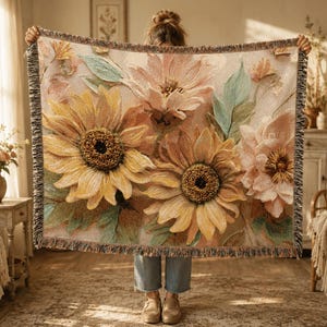 May include: A woven throw blanket featuring a large-scale floral pattern with prominent yellow sunflowers and soft pink blossoms set against a light cream background, finished with a decorative fringed edge on all four sides of the rectangular textile.