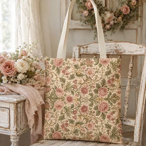May include: A rectangular fabric tote bag featuring a repeating pattern of pink roses and green foliage on a cream background. The bag has two sturdy, off-white canvas shoulder straps attached to the top edge, providing a simple, functional design.